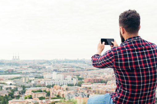 back view of a man taking a photo with his mobile phone of the city views from a rooftop, concept of freedom and technology, copy space for text
