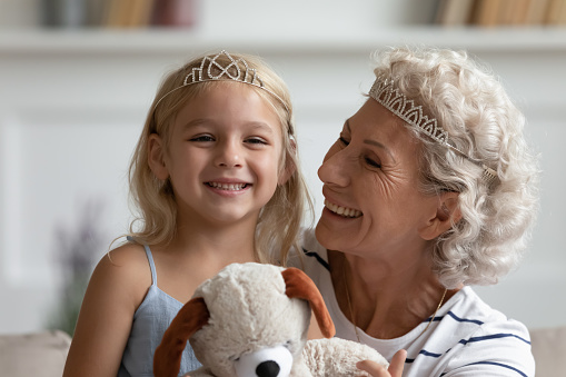 Queen and princess. Portrait of cute happy small child girl preschooler looking at camera cuddling and playing with beloved nanny or grandmother putting on crowns and creating fairy tale story game