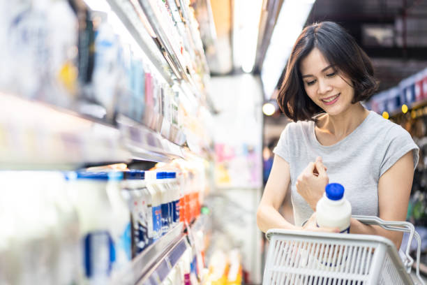 junge asiatische schöne frau hält lebensmittelkorb zu fuß im supermarkt. sie wählt das tägliche milchprodukt, das aus dem regal abgeholt wird. von der seite gesehen, während sie sich produkte anschaut. shopping-konzept. - color image food dairy product people stock-fotos und bilder