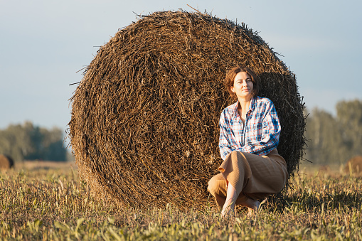 Romantic brunette woman in a plaid shirt and trousers  sitting on a round haystack in an amazing field, on background  forest. Warm colors of the sunset.