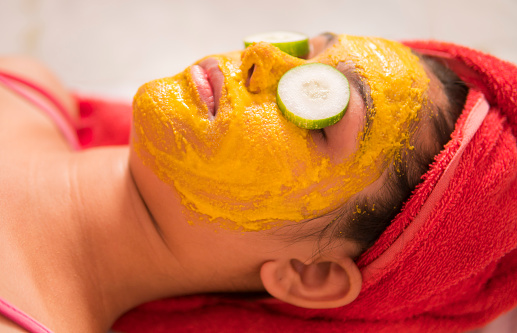Close-up image of woman applied beauty face mask with cucumber slice on the eyes and relaxing. She is wearing a red towel on the head and lying down.