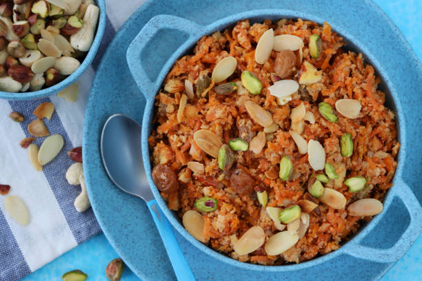 Stock photo showing grated carrot that has been cooked with water, milk and sugar and garnished with almonds. Traditional dessert severed at Indian festivals including Diwali and Holi. imagen tazón azul y plato con cuchara, gajar ka halwa (halwa de zanahoria), dulce budín de postre indio, tazón de zanahoria rallada endulzada y almendras junto a la toalla de té y tazón de nueces mixtas de almendra, anacardo y pistacho, vista elevada