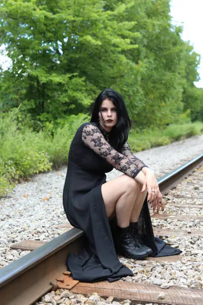 Photo of Spooky woman dressed in long black dress sitting on railroad tracks