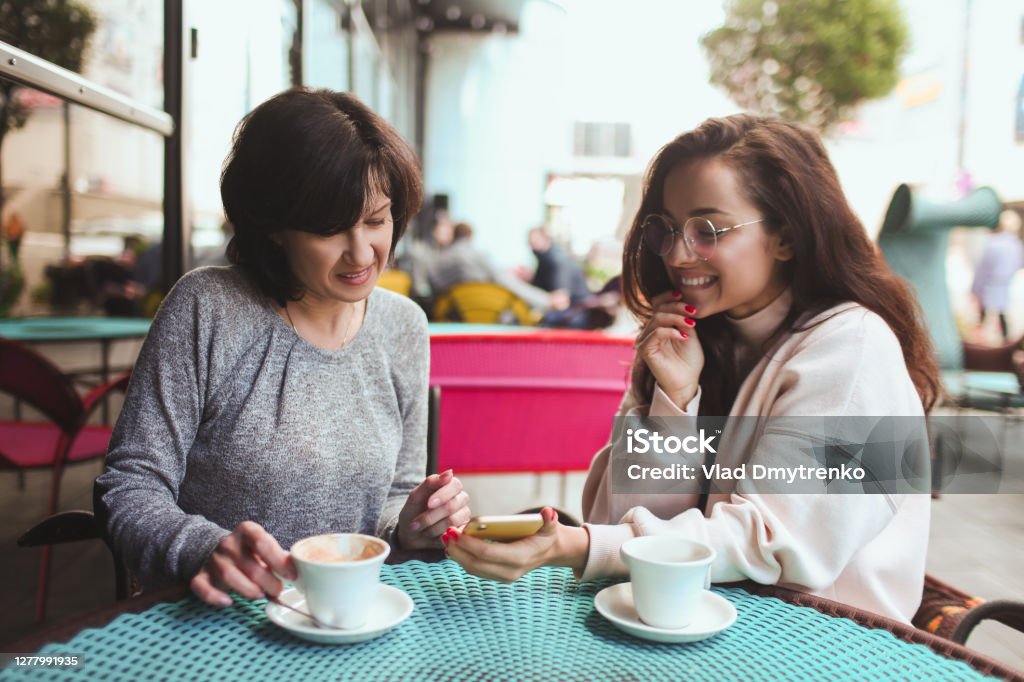 Mature mother and her young daughter sit together in cafe or restaurant. Girl hold smartphone and showing something on screen to her mom. Mature woman watch it. Drinking tea or coffee. Mature mother and her young daughter sit together in cafe or restaurant. Girl hold smartphone and showing something on screen to her mom. Mature woman watch it. Drinking tea or coffee Coffee - Drink Stock Photo