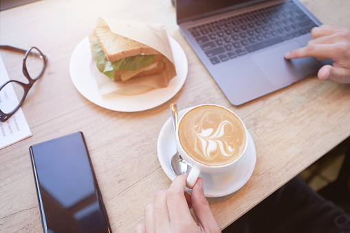 Top view of a freelancer or businessman using laptop, drinking coffee and having breakfast while working remotely in cafe. Business concept, distance work