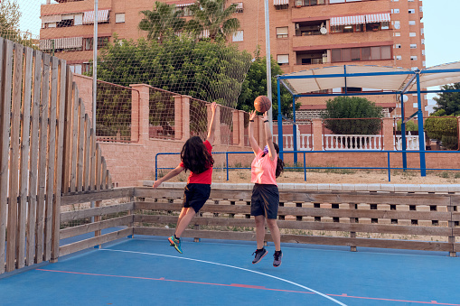 Two young girls play basketball on a city street court, one throwing and the other trying to block out, with the city buildings in the background.