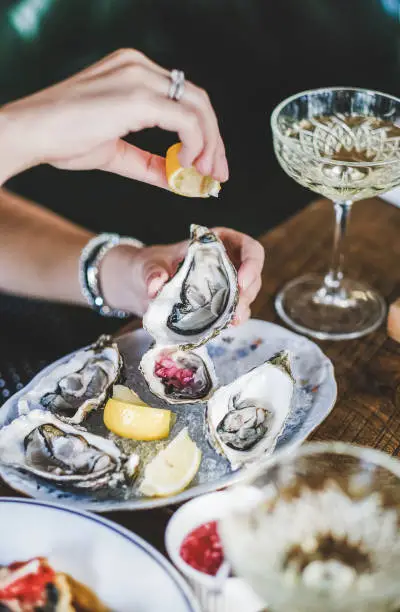 Hands of woman squeezing lemon juice to Irish oysters in plate with ice over glass of champagne in fish restaurant at background, selective focus. Seafood, French cuisine, fine dining concept