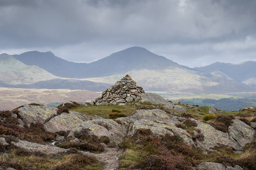 A shot of a rock pile on top of a hill in the English Lake District. In the background is a mountain range and a cloudy sky.