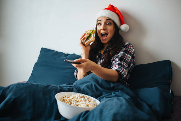 jeune belle femme dans le lit matinal à la maison. bonne gaie personne excitée regarder la télévision et utiliser la télécommande. période de noël ou du nouvel an. mangez du sandwich et du pop-corn. - freedom sandwich bread food photos et images de collection