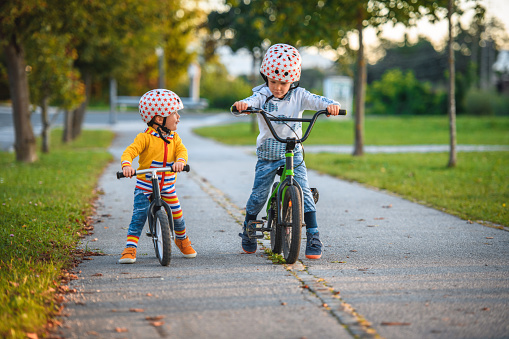 Two caucasian brothers with sports helmets driving bike and balance bike outdoors on a sunny day. They are supporting each other.