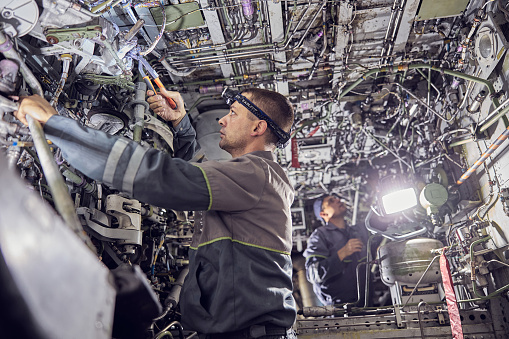 Side view portrait of two professional mechanics wearing overall uniform using professional aviation tools while standing inside aircraft