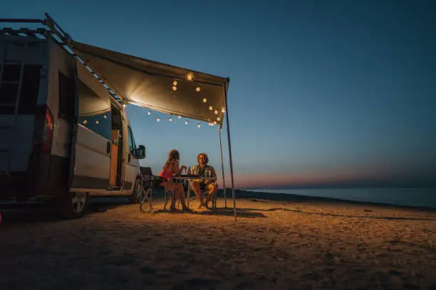 Young Caucasian heterosexual couple near the camper van parked near  the sea at sunset