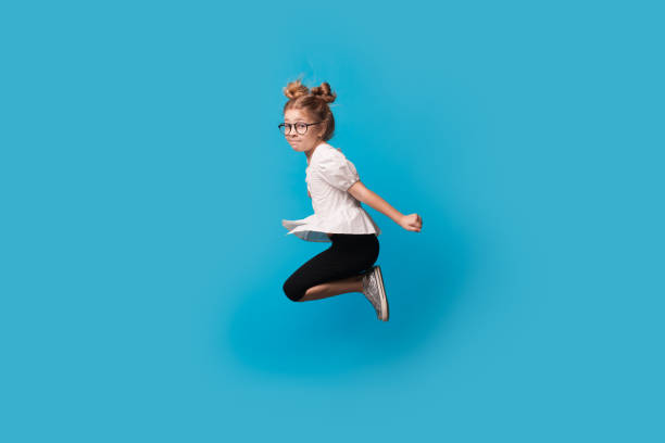 young girl with eyeglasses and blonde hair jumping on a blue studio wall - preschooler childhood outdoors cheerful imagens e fotografias de stock