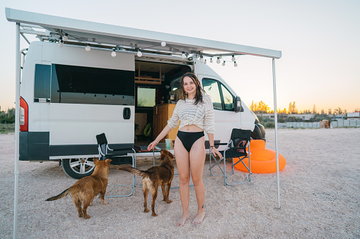 Young Caucasian woman  playing with dogs near the camper van parked near  the sea at sunset