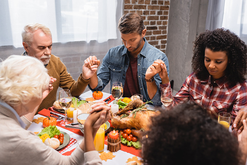 selective focus of multiethnic family with closed eyes holding hands during dinner on thanksgiving holiday
