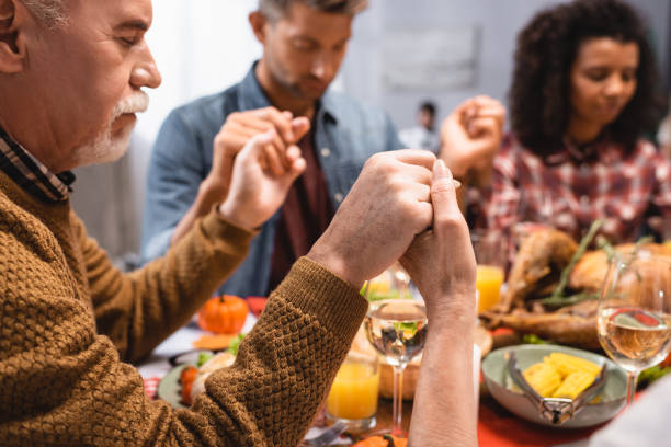 selective focus of senior man holding hands with multicultural family on thanksgiving selective focus of senior man holding hands with multicultural family on thanksgiving saying grace stock pictures, royalty-free photos & images