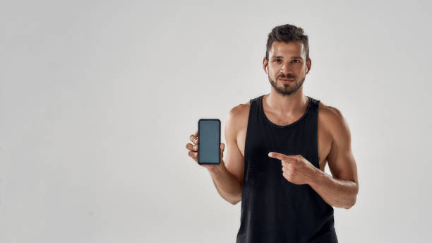 istock Handsome athletic man pointing at his smartphone with blank screen and looking at camera while standing in studio isolated over grey background, using fitness app 1278022154