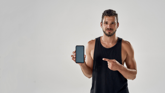 Handsome athletic man pointing at his smartphone with blank screen and looking at camera while standing in studio isolated over grey background, using fitness app. Sport and technology