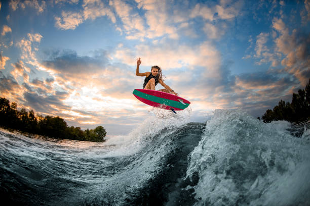 great view of woman jumping over big splashing wave on surf style wakeboard. great view of woman in swimsuit and vest actively jumping over big splashing wave on bright surf style wakeboard. surfing stock pictures, royalty-free photos & images