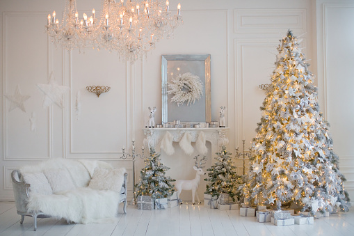 Cozy white Christmas interior room. Living room with Christmas tree, white sofa, crystal chandelier, fireplace and white and silver decorations