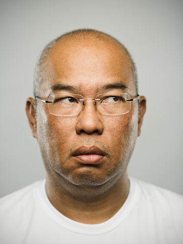 Close up portrait of asian mature adult man with blank expression looking to the side against gray white background. Vertical shot of real chinese man staring in studio with bald head and glasses. Photography from a DSLR camera. Sharp focus on eyes.