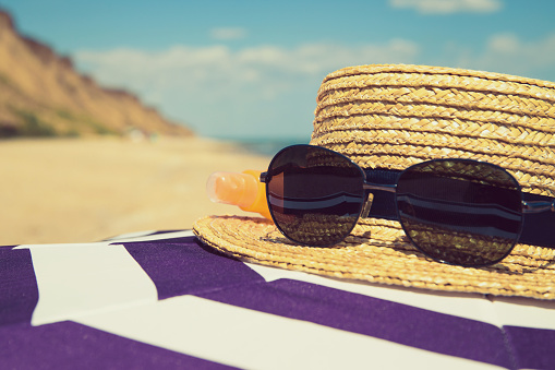 Striped  beach umbrella with straw hat, sunglasses and a bottle of sunscreen lotion on the sea shore with sand, water and vivid blue sky as a background. Summer vacation and travel concept. Image is with copy space