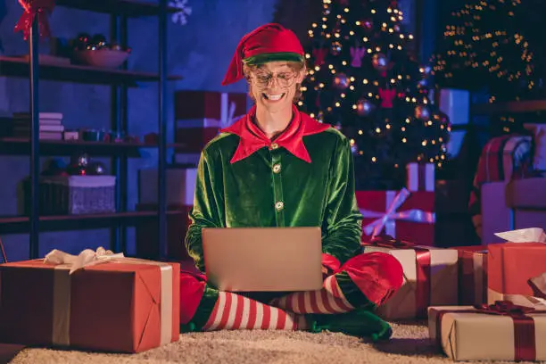 Full length photo of elf sit floor type greeting on laptop, wear green glasses cap in house indoors with evening x-mas decor lights