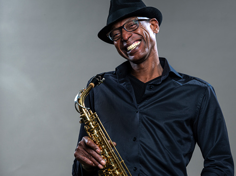 Musician with saxophone  against a grey backdrop. Man looking at camera with a smile, wearing a black hat and shirt