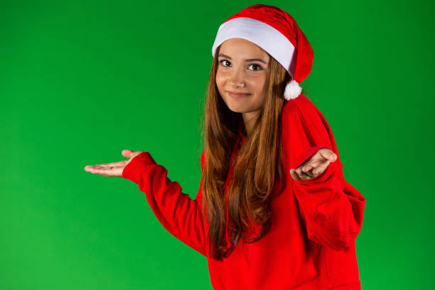 Beautiful caucasian woman in santa hat and red pullover showing something on empty hands, isolated over green background. High quality photo