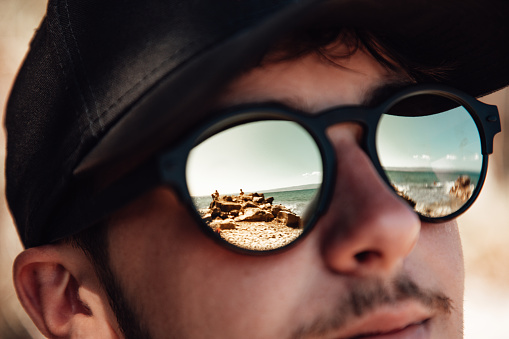 Young man wearing sunglasses mirroring the mediterranean croatian beach. Young man wearing a black cap and a black sunglasses. Selective Focus on reflections on sunglasses. Real People Millennial Generation Beach Vacation Lifestyle Concept. Dalmatia, Croatia, Europe.
