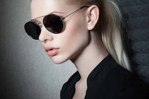 Closeup portrait of a sexy blonde female model with makeup and ponytail hairstyle, wearing black shirt and sunglasses.