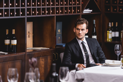 Selective focus of young man in suit looking away while sitting near glass of wine on table in restaurant