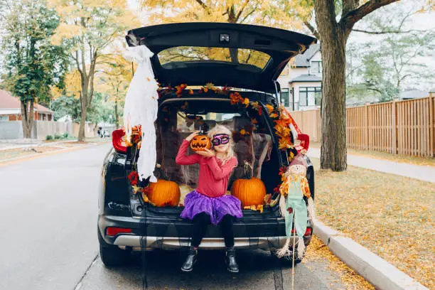 Photo of Trick or trunk. Child girl celebrating Halloween in trunk of car. Kid with red carved pumpkin celebrating traditional October holiday outdoors. Social distance during coronavirus covid-19.