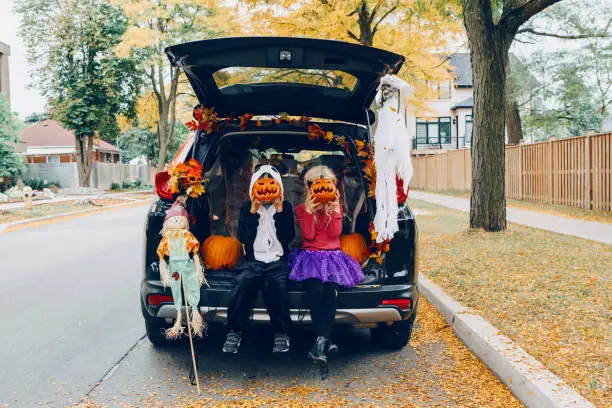 Photo of Trick or trunk. Children celebrating Halloween in trunk of car. Boy and girl with red pumpkins celebrating traditional October holiday outdoors. Social distance during coronavirus covid-19.