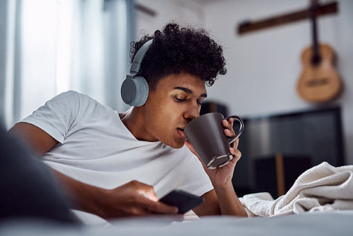 Shot of a young man having coffee and using a smartphone with headphones while relaxing on his bed