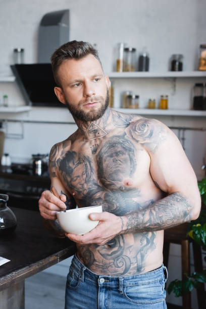istock muscular, tattooed man looking away while holding bowl with breakfast in kitchen 1278103452