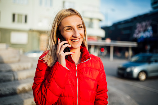 Happy Caucasian blonde woman using her smart phone outdoors, wearing a red jacket and smiling