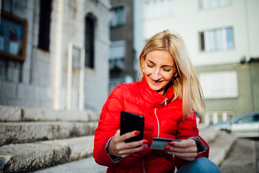 Beautiful Caucasian blonde woman wearing a red jacket, smiling and using her smart phone, holding a credit card