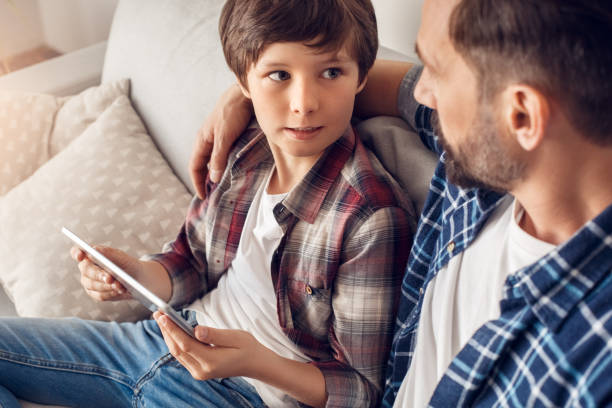 お父さんと小さな息子は、お互いを見て、デジタルタブレットで男の子を抱きしめるソファのお父さんの上に座って深刻なクローズアップ - digital tablet family father offspring ストックフォトと画像