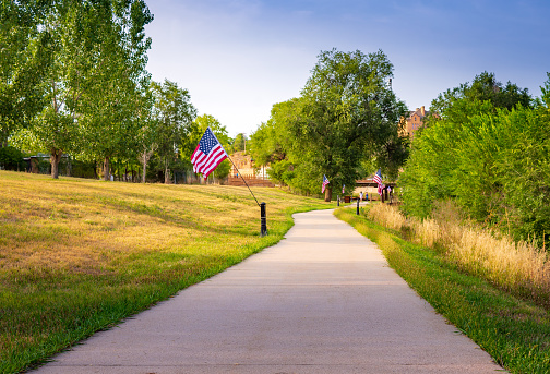 A cement sidewalk winding though a tree lined park that has multiple American flags lining the walk.