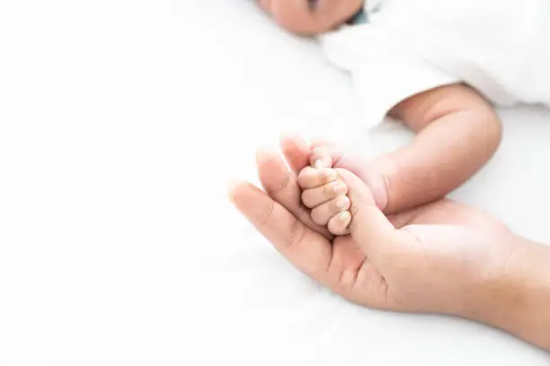 Photo of Newborn baby holding motherâs finder while sleeping on white bed. Mother takes care and comforting for her baby