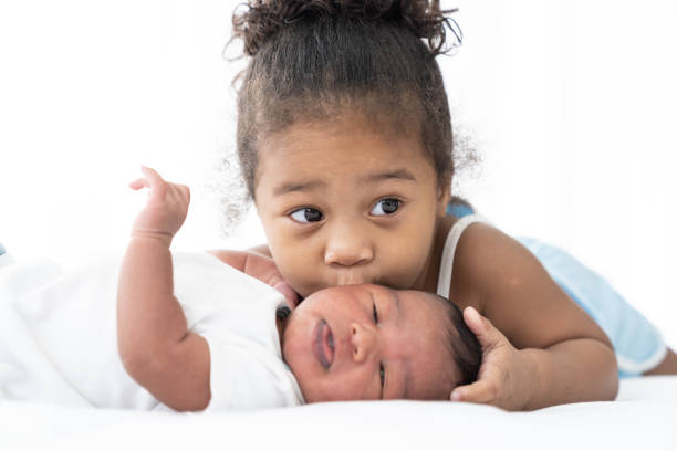 menina afro-americana fofa beijando em bochecha de bebê recém-nascido na cama branca em casa. menina cuida de bebê com gentil - 18 a 23 meses - fotografias e filmes do acervo