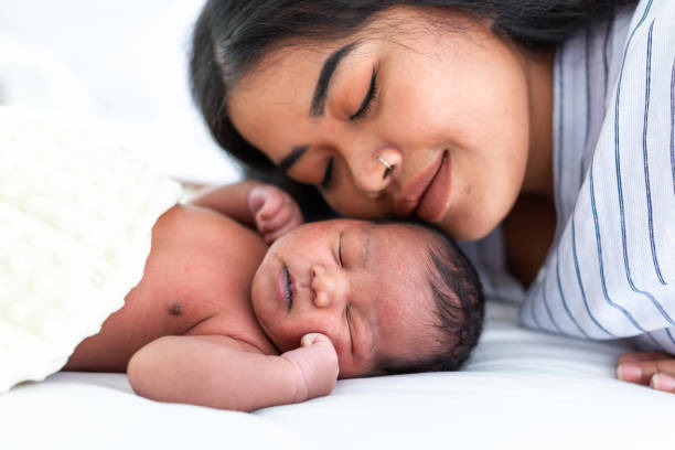 afroamerikanische mutter umarmt und küsst ihr neugeborenes baby auf weißem bett. nahaufnahme des säuglings mit der jungen mutter - new mother stock-fotos und bilder