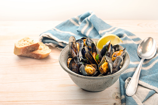 Bowl with mussels, lemon and herbs, a blue towel, spoon and bread on a light wooden table, copy space, selected focus, narrow depth of field
