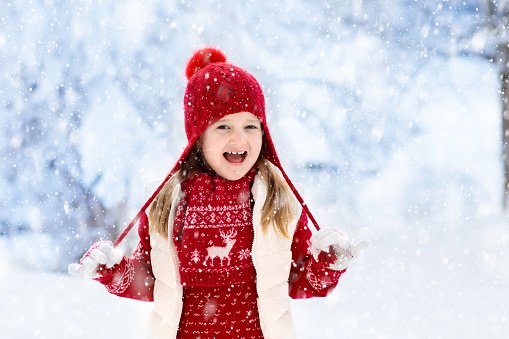 Child in red hat playing in snow on Christmas vacation. Winter outdoor fun. Kids play in snowy park on Xmas eve. Little girl in knitted sweater, scarf and mittens with Christmas decoration.