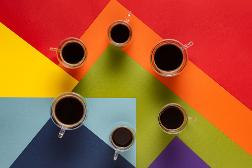Circle made of six glass round cups with fresh black espresso and Americano coffee. Colorful abstract background with geometric pattern in rainbow colors and with paper texture. Directly above view.
