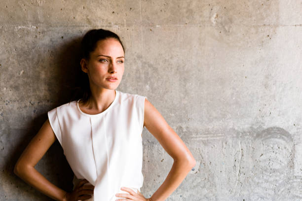 Thoughtful young woman looking away against gray concrete wall. Beautiful female is standing with hands on hip. She is in casuals.