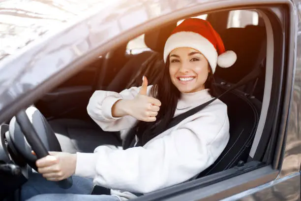 Happy New Year and Merry Christmas! A woman sits in a car, she is dressed in a red Santaclaus hat and shows a thumb up.