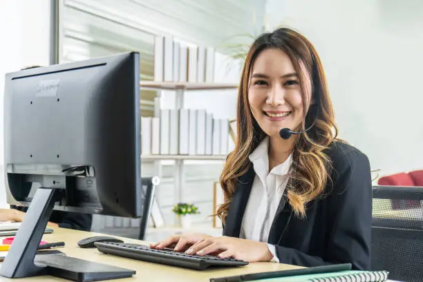 Smiling friendly asian female call-center agent with headset working on support hotline in the office