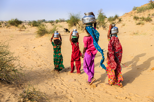 Indian little girls crossing sand dunes and carrying on their heads water from local well, Thar Desert, Rajasthan, India. Rajasthani women and children often walk long distances through the desert to bring back jugs of water that they carry on their heads.